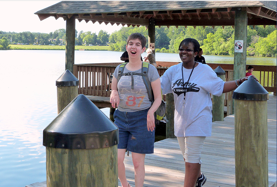 A person supported by a staff walking by a lake